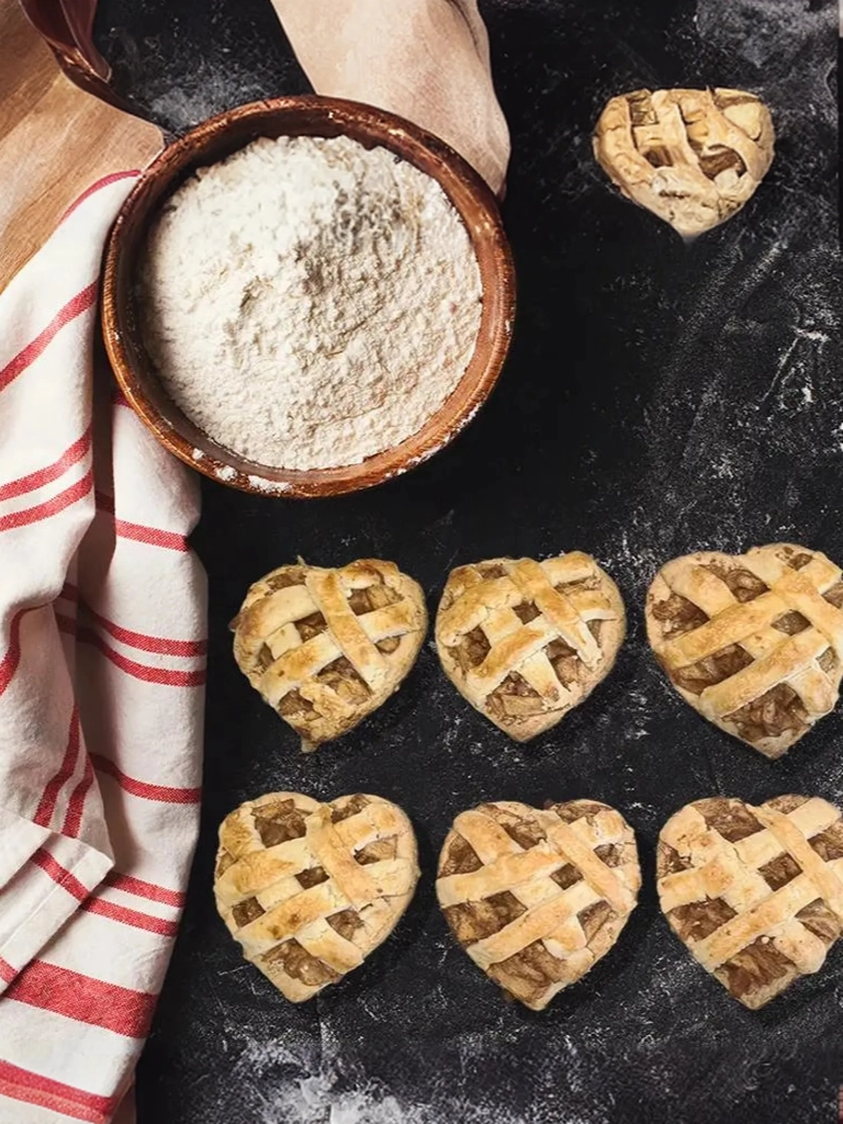 Zes hartvormige appeltaart koekjes met een raster van deeg, gepresenteerd op een met bloem bestoven ondergrond naast een houten kom met bloem en een rood-wit gestreepte doek.