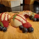 Witte chocoladebollen gevuld met room en bosvruchten, gegarneerd met frambozen, blauwe bessen en een rode saus, geserveerd op een houten plank.