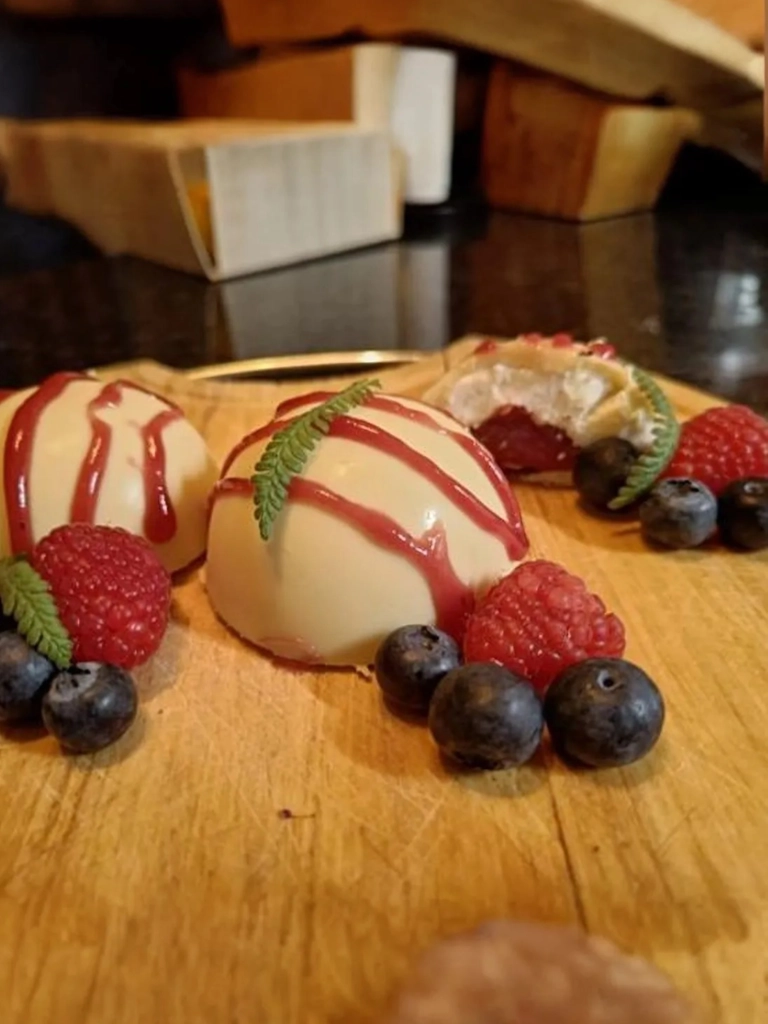 Witte chocoladebollen gevuld met room en bosvruchten, gegarneerd met frambozen, blauwe bessen en een rode saus, geserveerd op een houten plank.