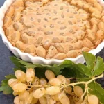 Een versgebakken clafoutis met sappige druiven in een witte ovenschaal, geserveerd op een donkere ondergrond, vergezeld door een tros verse witte druiven met bladeren.