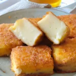 Een bord met goudbruin gebakken blokjes leche frita, een traditioneel Spaans dessert, gegarneerd met suiker en kaneel, met een glas thee op de achtergrond.