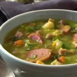 Een dampende kom traditionele erwtensoep met stukjes rookworst, spek en groenten, gegarneerd met verse kruiden, geserveerd met vers brood op de achtergrond.