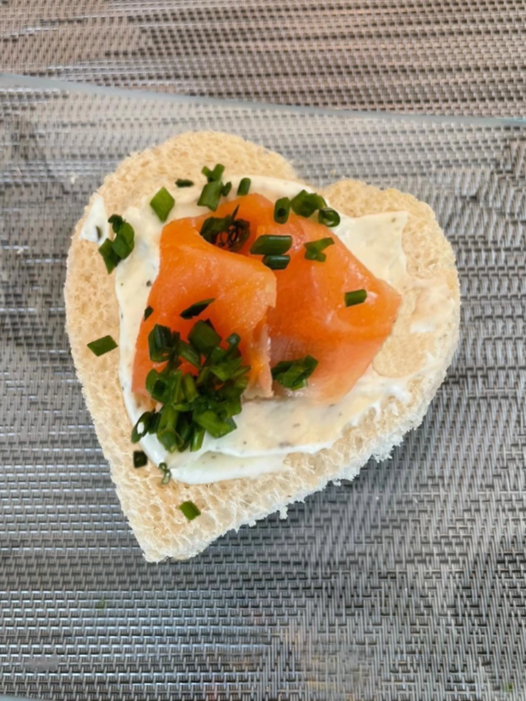 Een hartvormige toast belegd met een laagje roomkaas, tonijn, gerookte zalm en vers gesneden bieslook, gepresenteerd op een geweven ondergrond.