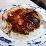 Babi pangang, plakjes gemarineerd en geroosterd varkensvlees geserveerd op een bedje van witte rijst, gepresenteerd op een decoratief wit-blauw bord