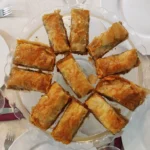 Een schaal met goudbruine stukken Griekse baklava, bestaande uit lagen filodeeg, walnoten en honing, gepresenteerd op een eettafel met wit linnen en servies.
