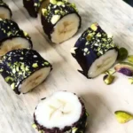 Plakjes banaan omhuld met pure chocolade en bestrooid met fijngehakte pistachenoten, gepresenteerd op een houten plank.