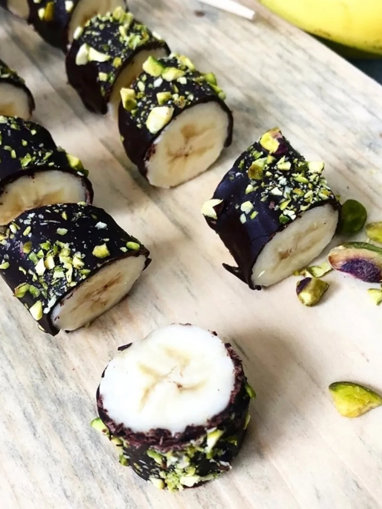 Plakjes banaan omhuld met pure chocolade en bestrooid met fijngehakte pistachenoten, gepresenteerd op een houten plank.