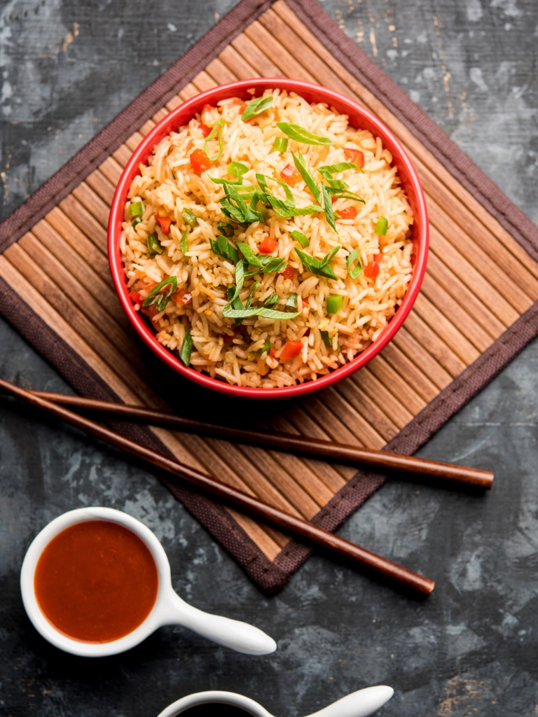 Een kom met Chinese nasi, gegarneerd met gesneden lente-uitjes en rode paprika, met saus en eetstokjes aan de zijkant.