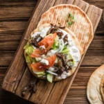 Een bord met traditionele Griekse gyros, geserveerd met pitabrood, tzatziki, gesneden groenten zoals tomaat en ui, en een schijfje citroen.