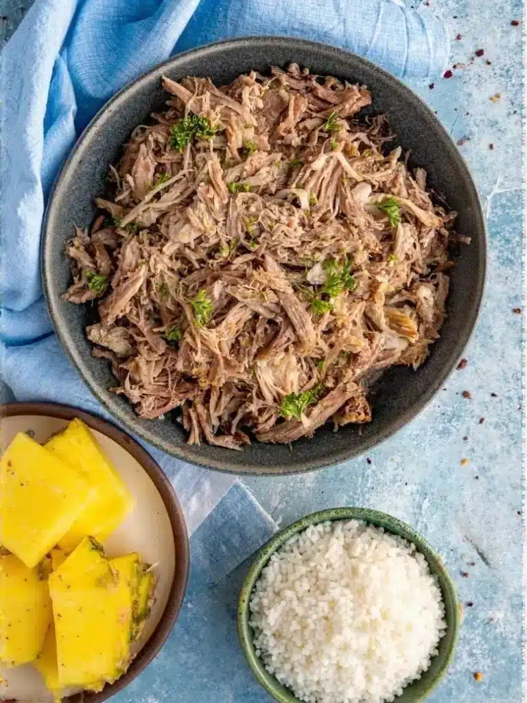 Hawaiiaanse Kalua pork, langzaam gegaard en gekruid varkensvlees, geserveerd op een bed van witte rijst met een schijfje kerstomaat, gepresenteerd op een lichtblauw bord.
