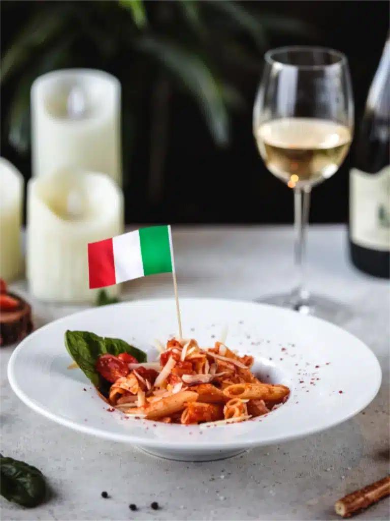 en elegant gepresenteerd bord met penne in een rijke tomatensaus, gegarneerd met basilicum en geraspte kaas, met een Italiaanse vlag in het midden. Op de achtergrond kaarsen, een glas witte wijn en een wijnfles voor een authentieke sfeer.