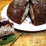 Maulwurfkuchen, een chocoladekoepeltaart gevuld met room en banaan, bestrooid met poedersuiker en gegarneerd met zwarte bessen en een takje munt, geserveerd op een wit bord.