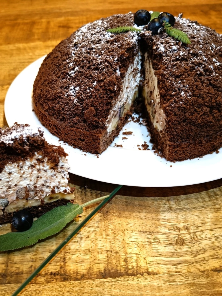 Maulwurfkuchen, een chocoladekoepeltaart gevuld met room en banaan, bestrooid met poedersuiker en gegarneerd met zwarte bessen en een takje munt, geserveerd op een wit bord.