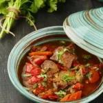 Muckalica, een Servisch stoofgerecht met mals varkensvlees, paprika, tomaten en wortels in een hartige saus, geserveerd in een decoratieve aardewerken schaal met een deksel, met verse peterselie op de achtergrond.