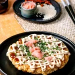 Een okonomiyaki, een Japanse hartige pannenkoek, belegd met mayonaise, sesamzaadjes, lente-ui en ingelegde zalm. Geserveerd met een klein kommetje saus en chili's.