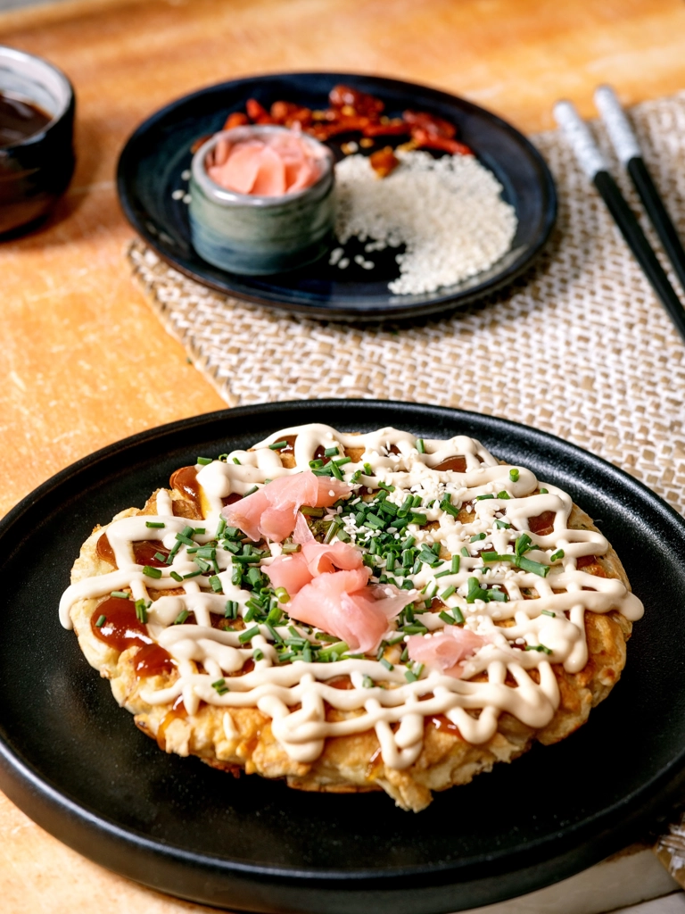 Een okonomiyaki, een Japanse hartige pannenkoek, belegd met mayonaise, sesamzaadjes, lente-ui en ingelegde zalm. Geserveerd met een klein kommetje saus en chili's.