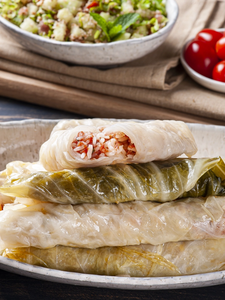Een schaal met Sarma, traditionele koolrolletjes gevuld met rijst en gehakt, waarvan één opengesneden is om de vulling te tonen, geserveerd met een salade en een schaaltje cherrytomaten op de achtergrond.