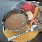 Een kom Servische goulashsoep geserveerd in een zwarte kom met witte patronen, vergezeld door een lepel op een houten plank, en groenten zoals paprika, wortel, ui en knoflook op de achtergrond.