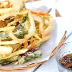 Een schaal met gefrituurde groenten in tempura-beslag, geserveerd met een klein kommetje sojasaus met sesamzaadjes.