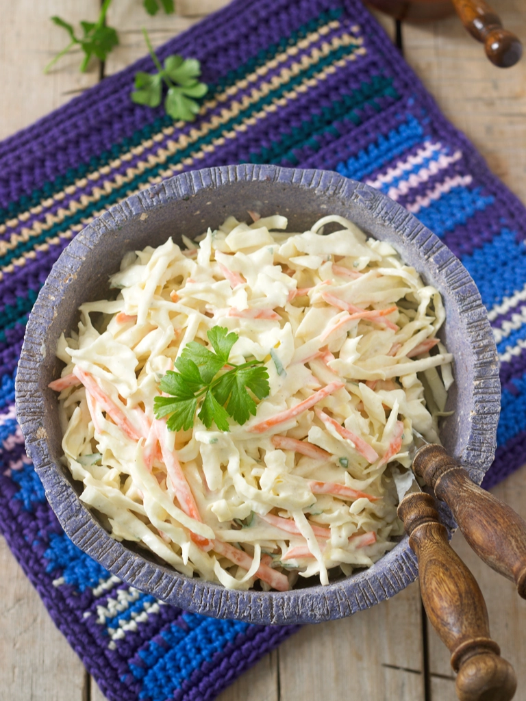 Een kom coleslaw gemaakt van fijngesneden kool, gemengd met een romige dressing en gegarneerd met een takje peterselie, geserveerd in een rustieke schaal op een kleurrijke gehaakte onderzetter.
