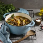 Een blauwe braadpan gevuld met choirinó stifádo, een Griekse stoofschotel met varkensvlees, aardappelen en groenten, geserveerd met een houten lepel en verse kruiden op de achtergrond.