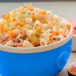 Een blauwe kom gevuld met Hawaiiaanse macaroni-salade, bestaande uit pasta, blokjes wortel, ananas, bleekselderij, en een romige dressing