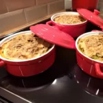 Drie rode ovenschalen met kipgratin, afgedekt met stokbrood en gesmolten kaas, klaar om te serveren, gepresenteerd op een kookplaat in een moderne keuken.