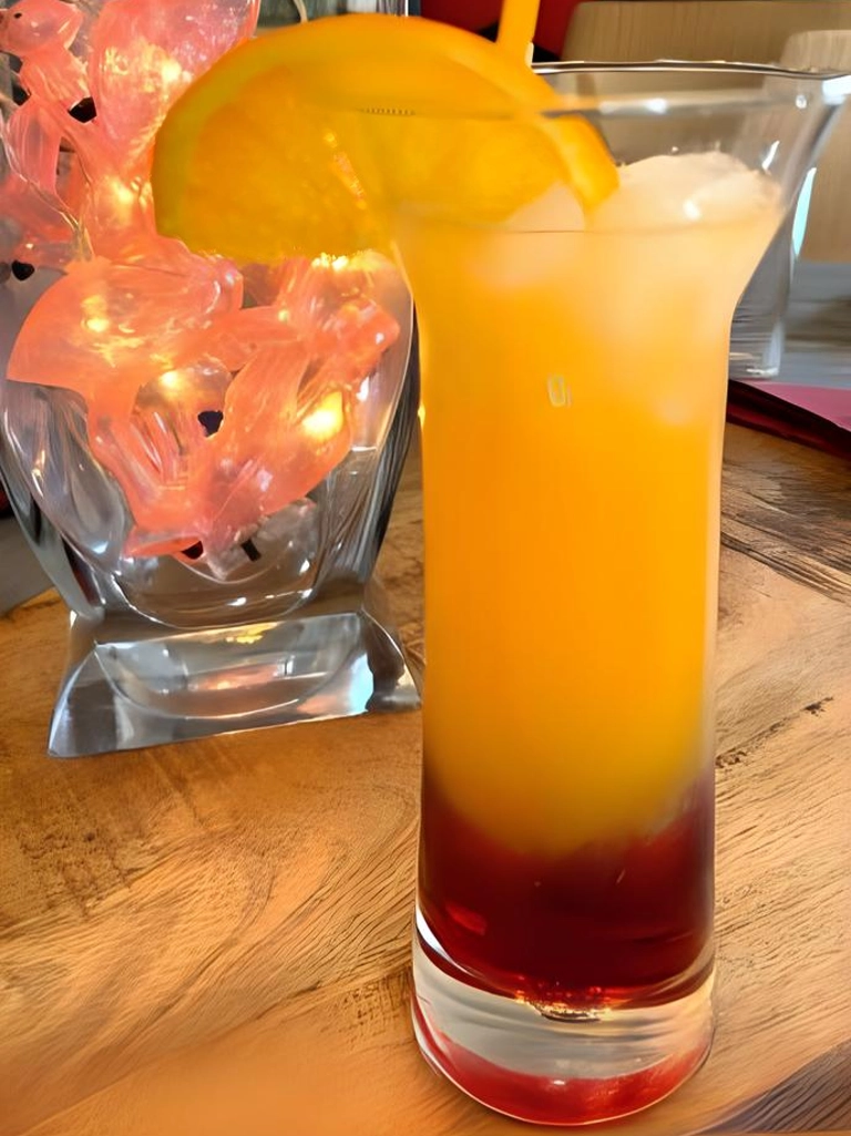 Een glas met een Sex on the Beach cocktail, met lagen van rood grenadine, sinaasappelsap en ijsblokjes, gegarneerd met een schijfje sinaasappel, op een houten tafel met sfeerverlichting op de achtergrond.