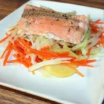 Een bord met gestoomde zalmfilet op een bedje van julienne gesneden wortels, prei en plakjes aardappel, gegarneerd met peper.