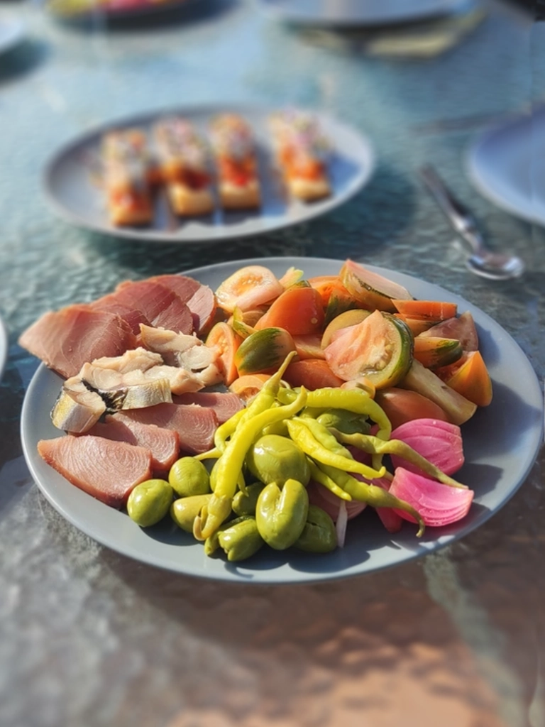 Mediterrane zomersalade met zee en streekproducten op een bord dat op een glazen tafel staat. Je ziet ondermeer groene olijven, groen pepertjes, tomaatjes en uien