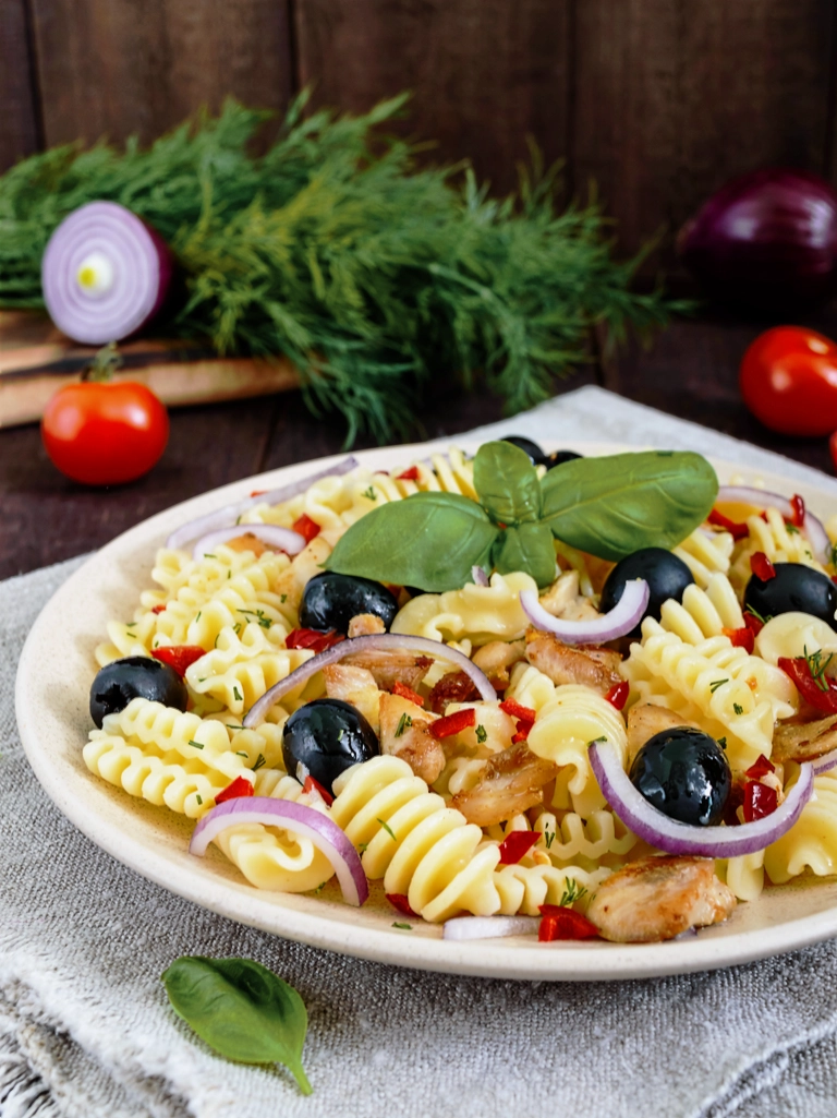 Pasta met tonijn en olijven op een licht bord met daaronder een grof geweven doek. De olijven zijn glimmen op het gerecht geplaatst en bovenop licht een takje basilicum